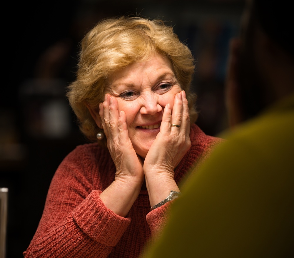 Vrouw zit met haar handen op haar wangen lachend te kijken naar de gesprekspartner tegenover haar.