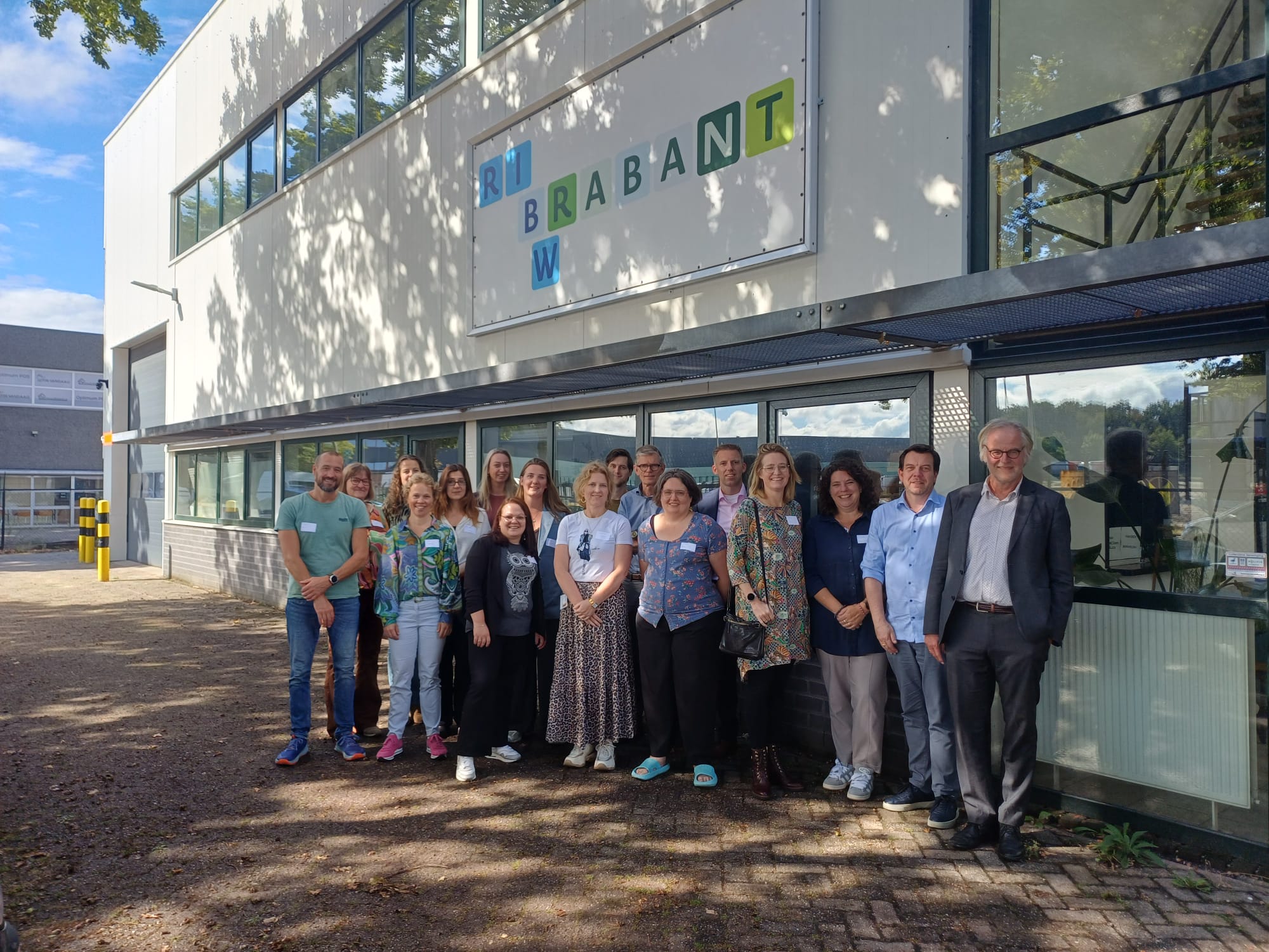 Delegatie van mannen en vrouwen van de ministeries van VWS en SZW samen met deelnemers en ervaringsdeskundigen in de schaduw van de bomen voor het pand van KREW op de Ledeboerstraat.