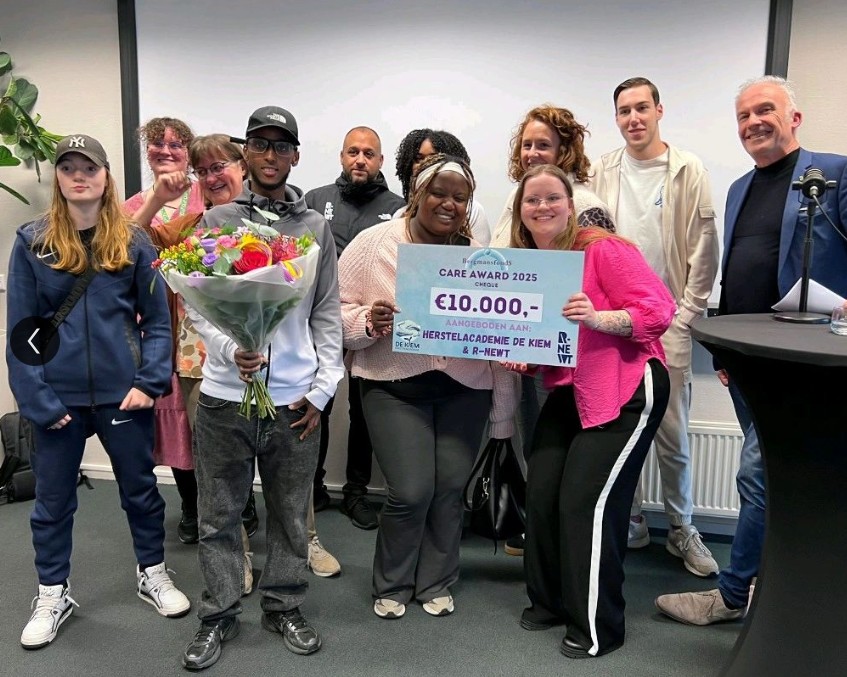Groep jongeren staat met bloemen en een cheque met daarop 10.000 euro naast de juryvoorzitter, trots dat ze de Care Award hebben gewonnen voor project Van Inzicht naar Impact van Herstelacademie De Kiem.